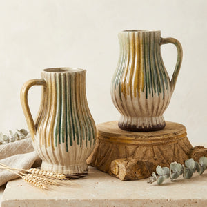 Two ribbed ceramic jugs with green and yellow drips, on a rustic surface with wheat and eucalyptus.