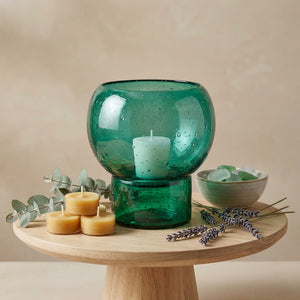 Green bubble glass vase with white candle, eucalyptus, lavender, and tealights on a wooden stand.