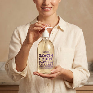 Smiling person holds a clear bottle of Marseille liquid soap with purple text on a beige background.