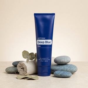 Blue tube labeled "Deep Blue Rub" surrounded by gray stones, eucalyptus leaves, and a rolled cloth.