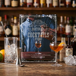 Cocktail book with drink, shaker, bitters bottles, and citrus on a wooden bar counter.