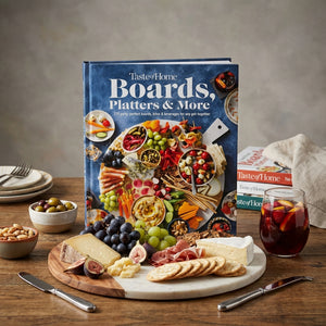 Cookbook titled "Boards, Platters & More" beside a cheese board with meats and fruits on a wooden table.