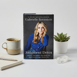 Book titled "Judgment Detox" with a woman on the cover, next to a plant, mug, and notebook.