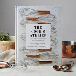 Open cookbook from The Cook’s Atelier by Abrams showing a colorful plated dish and kitchen utensils on a rustic table.
