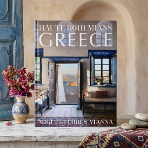 Book titled "Haute Bohemians Greece" on a table with a vase of dried flowers and a patterned rug.
