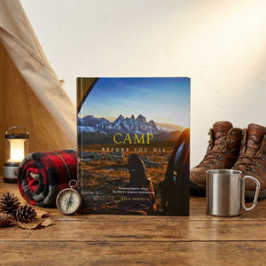 Camping book on a wooden table with boots, a metal mug, lantern, compass, and red plaid blanket.