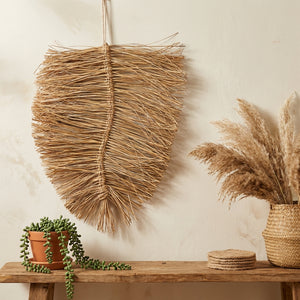 Woven leaf wall decor with pampas grass, potted succulent, and coasters on wooden bench. Neutral tones.