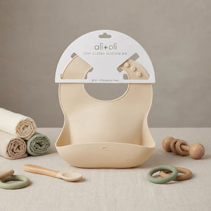 Beige silicone bib with packaging, surrounded by rolled cloths, wooden spoon, and teething rings.